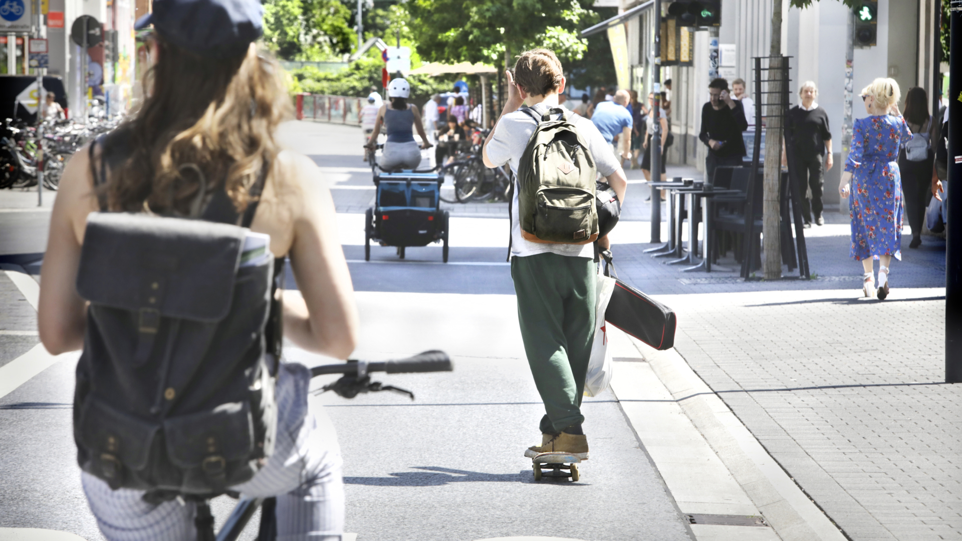 2025-10-27-Zinsereck 11 Eine belebte Stadtstraße bei Sonnenschein. Eine Frau mit Rucksack fährt auf einem Fahrrad, während ein junger Mann mit Rucksack und Skateboard vorausfährt. Weitere Menschen gehen oder fahren Fahrrad im Hintergrund.